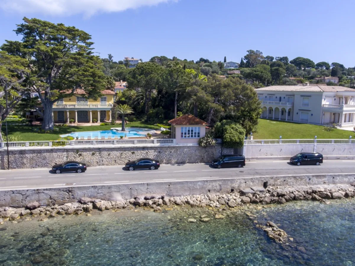 Flotte de véhicules de luxe Mercedes-Benz roulant sur une route côtière de la Côte d’Azur, devant des villas prestigieuses. Service de transfert VIP avec chauffeur privé pour un voyage exclusif et élégant.