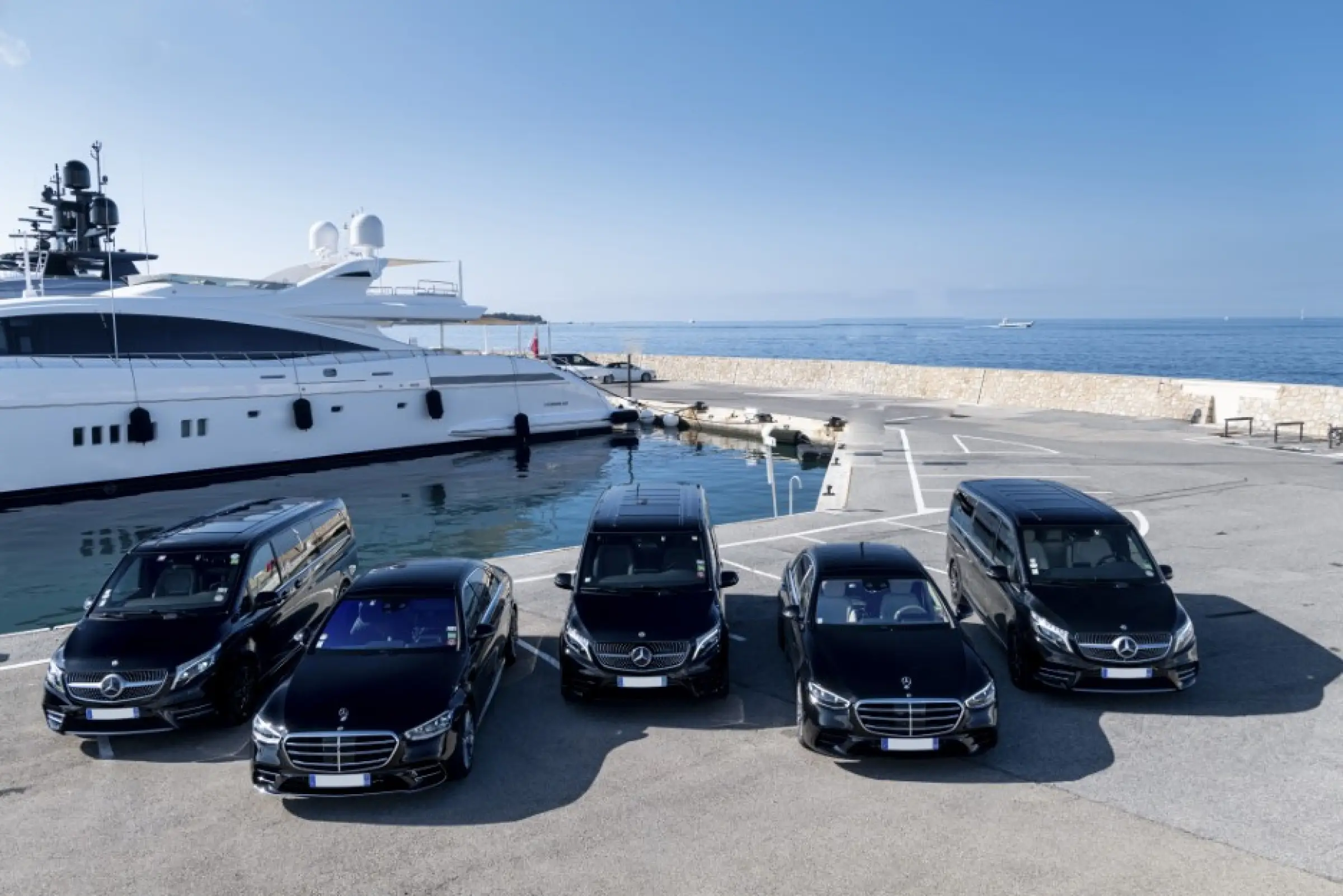 Flotte de véhicules de luxe Mercedes-Benz stationnée près d’un yacht sur la Côte d’Azur. Service de transfert VIP avec chauffeur privé pour un transport exclusif et haut de gamme.