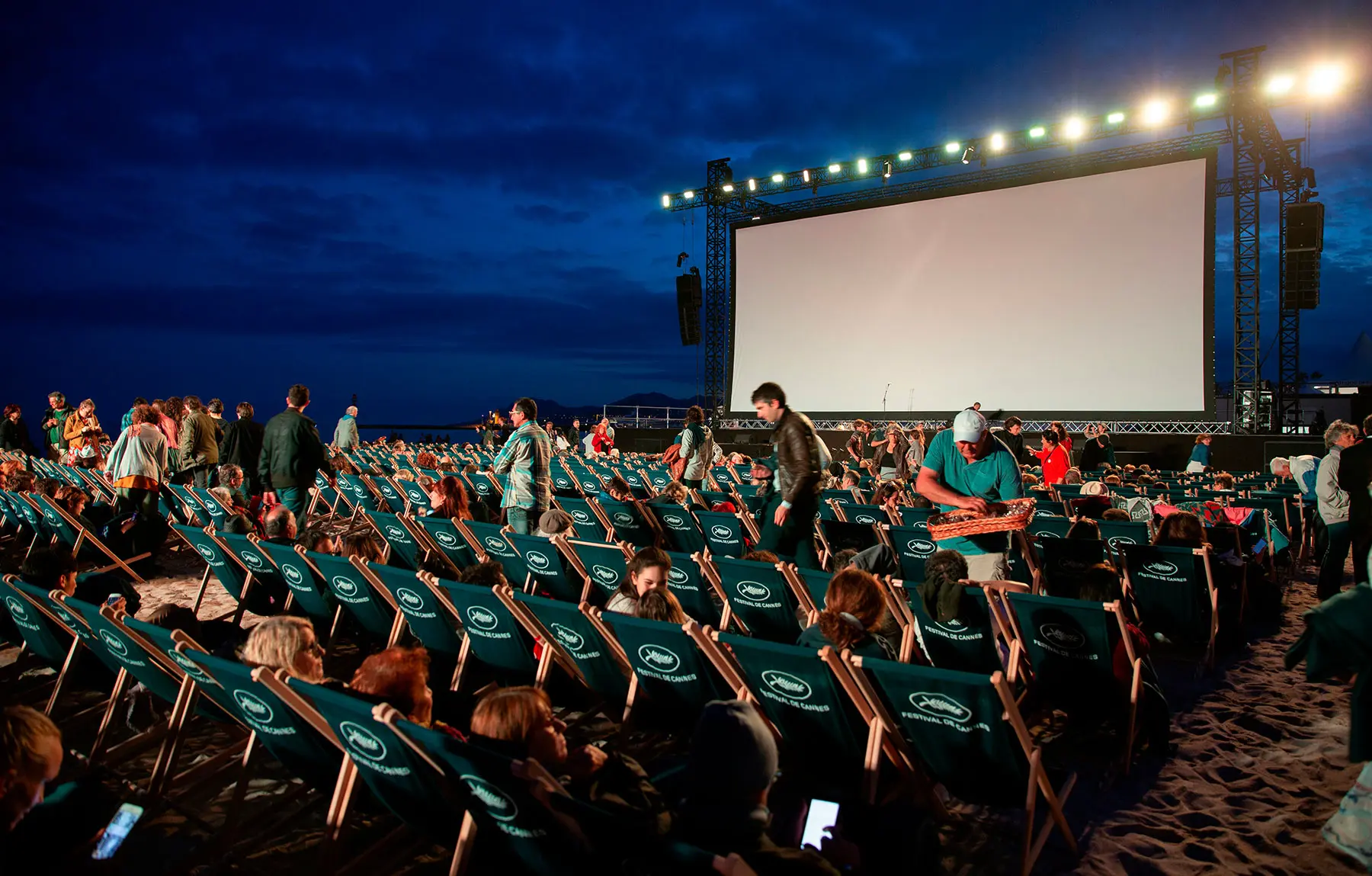 Open-air film screening on the beach, with a large crowd seated on “Festival de Cannes” deckchairs, just moments before the movie begins.