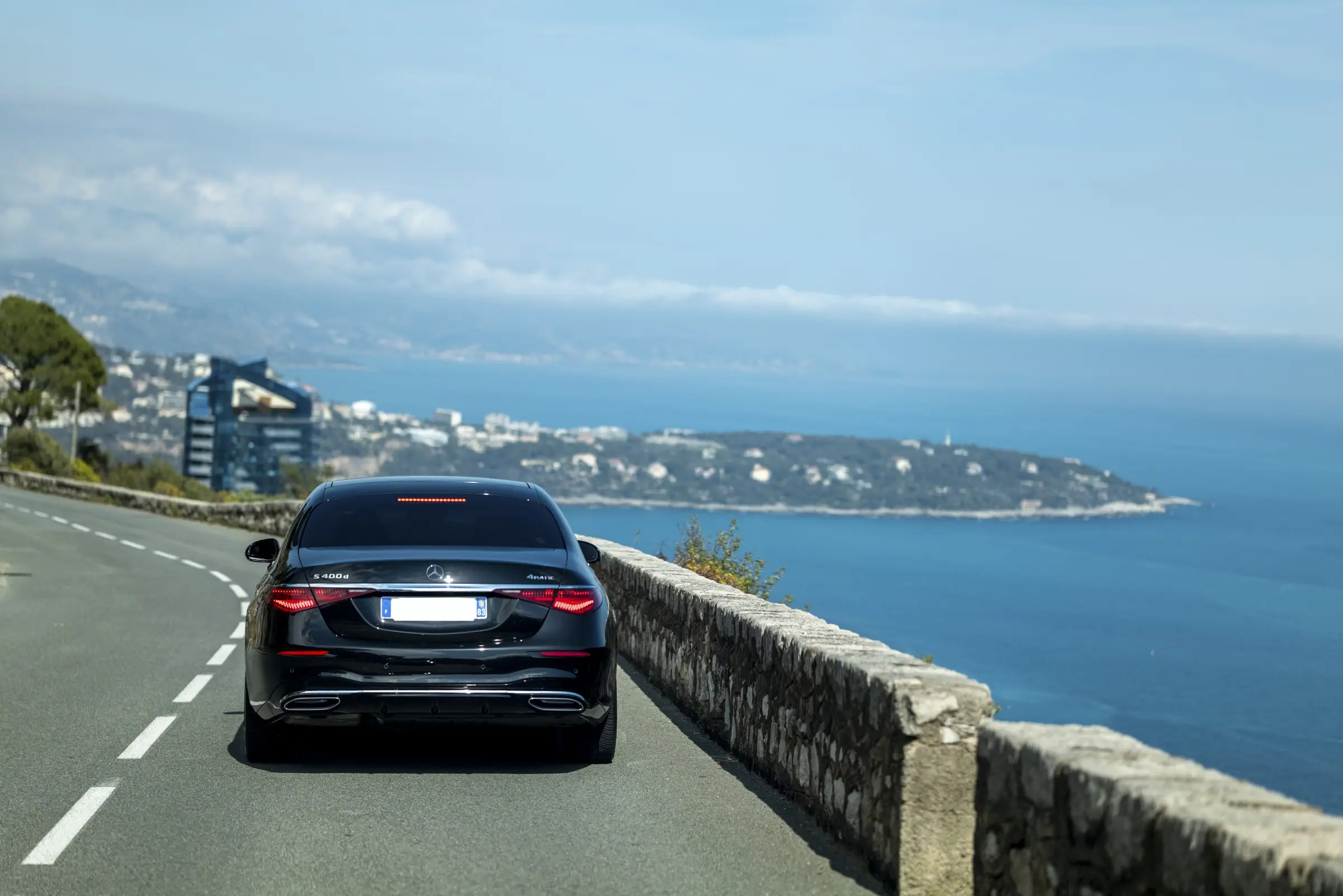 Rear view of a Mercedes S-Class luxury sedan driving along a scenic road overlooking the Mediterranean, with views of the French Riviera coastline and hills in the background. An ideal setting for a private chauffeur excursion. 