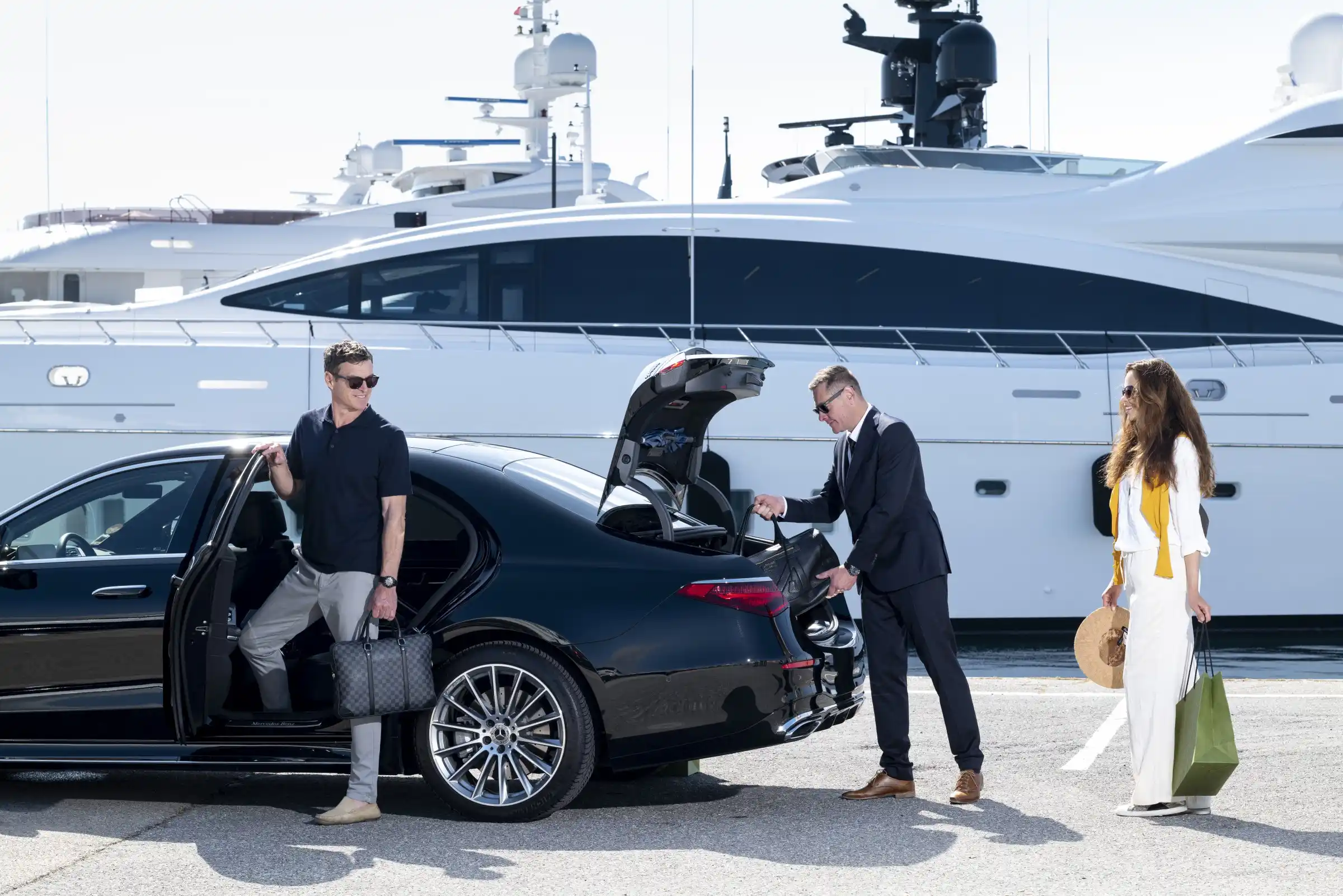 An elegant couple steps out of a black Mercedes S-Class while a chauffeur in a suit hands them their luggage in front of a yacht-filled marina. A VIP transfer scene between luxury land and sea transport, typical of the French Riviera. 