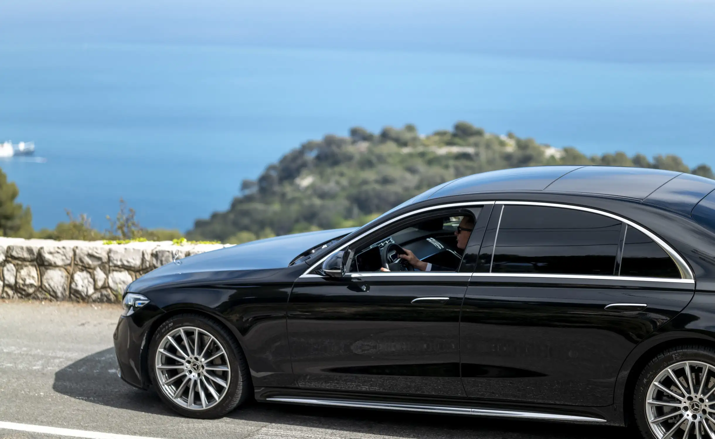 Chauffeur privé au volant d’une berline de luxe Mercedes Classe S sur une route panoramique surplombant la Méditerranée, en direction de Saint-Tropez. Service haut de gamme, confort, élégance et discrétion assurés pour une excursion sur la Côte d’Azur.