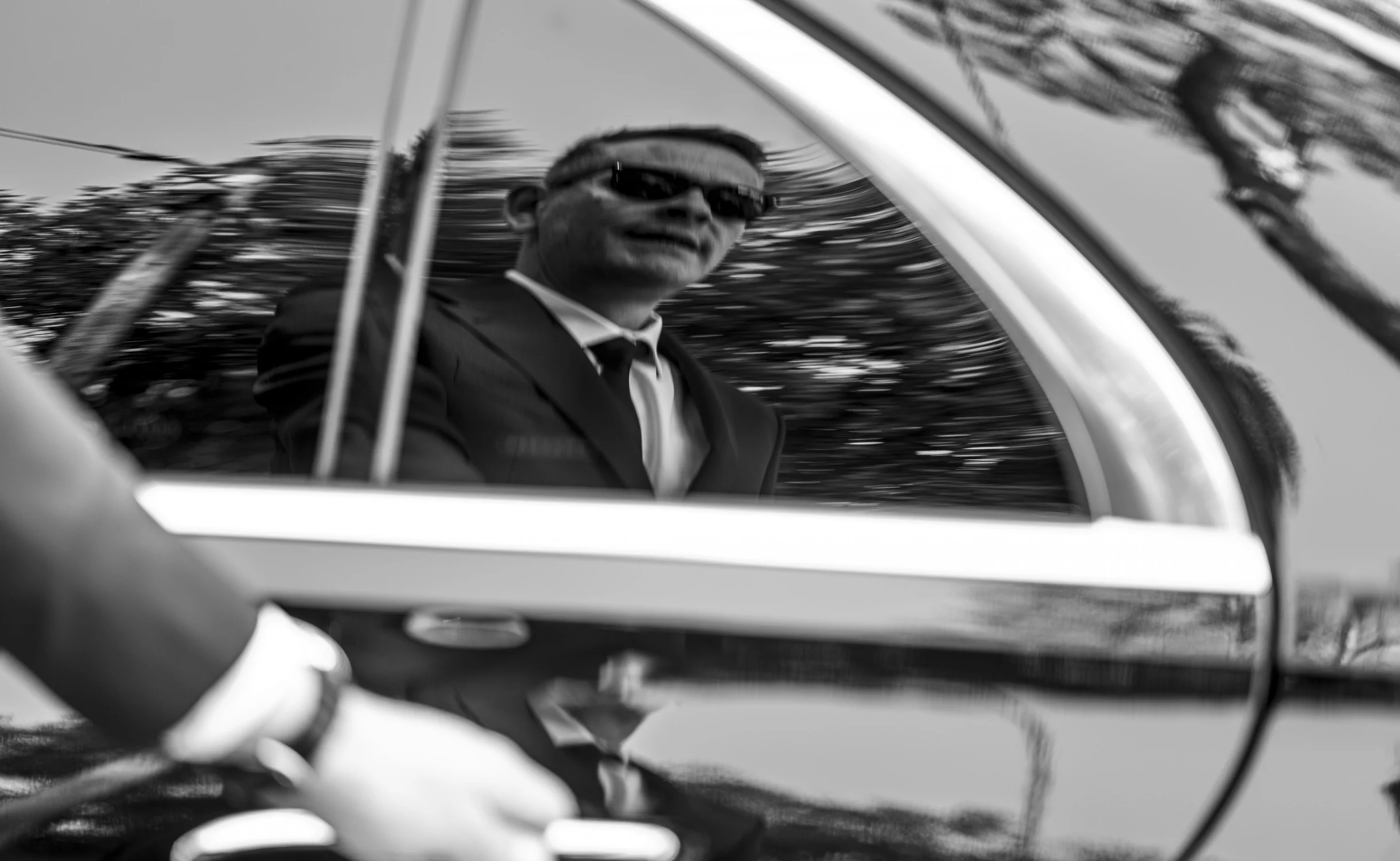 Vue en noir et blanc du reflet d’un chauffeur privé en costume et lunettes de soleil, visible sur la surface brillante d’une portière de voiture. Un cliché élégant et symbolique de discrétion, de professionnalisme et de service haut de gamme personnalisé à Cannes.