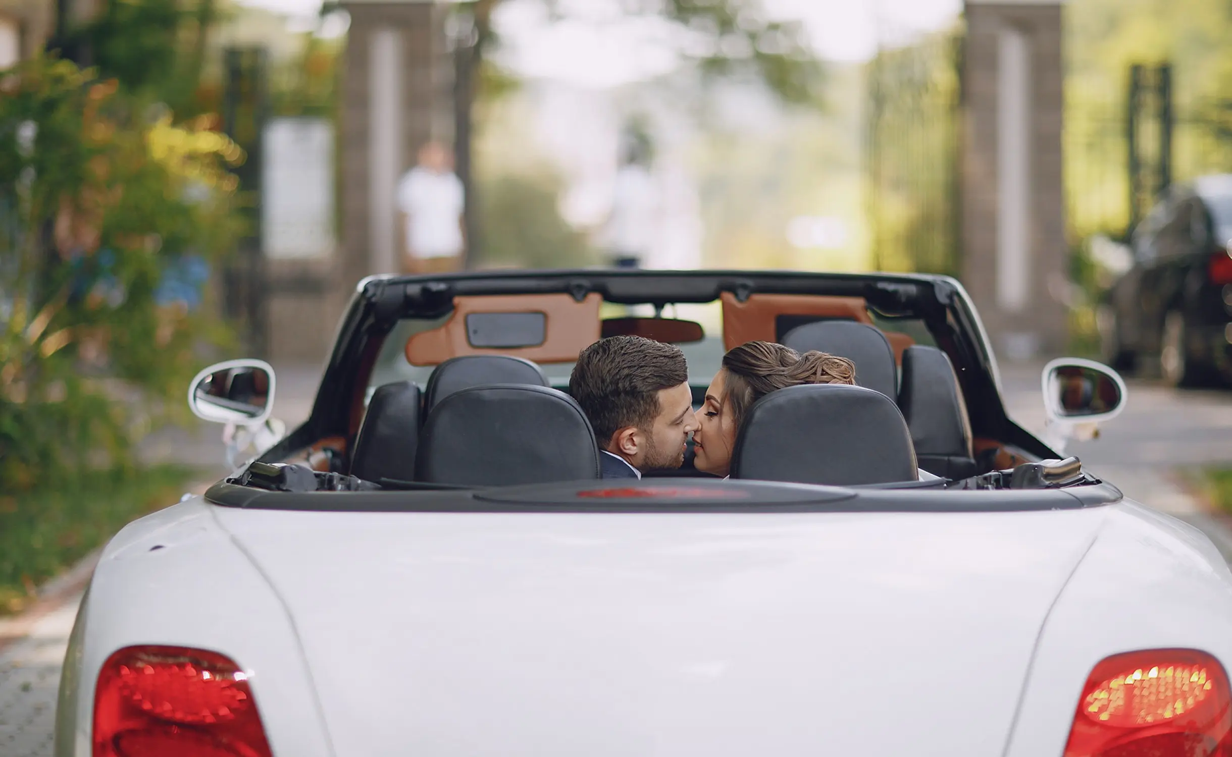Couple de jeunes mariés échangeant un baiser à l’arrière d’une voiture décapotable blanche de luxe – moment romantique d’un transport de mariage premium.