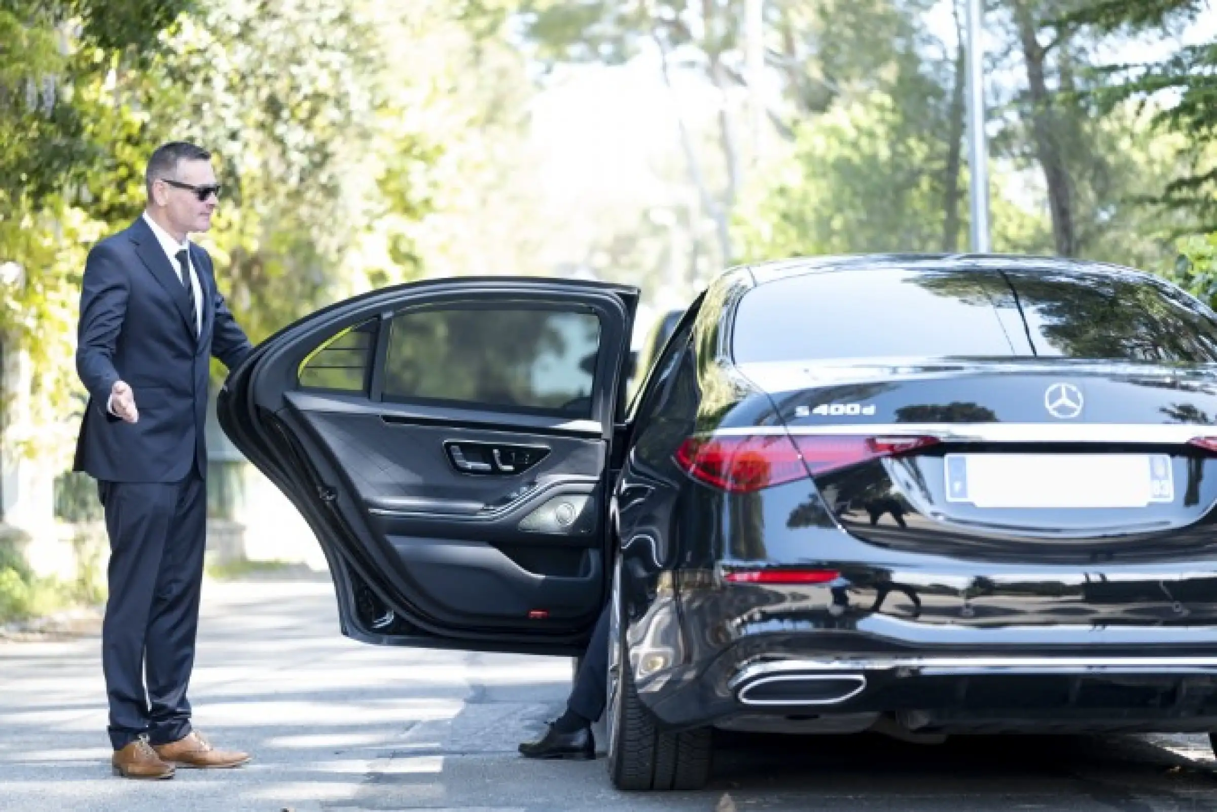 A professional chauffeur in a suit opens the rear door of a black Mercedes S 400d, ready to welcome a passenger in a secure and elegant setting surrounded by greenery.