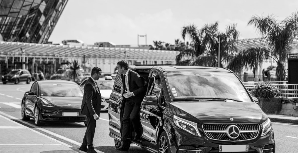 Un agent d’accueil en costume échange avec un client d’affaires descendant d’un van Mercedes classe V, devant un terminal d’aéroport. Le service de transfert illustre le haut niveau de confort et de discrétion offert.