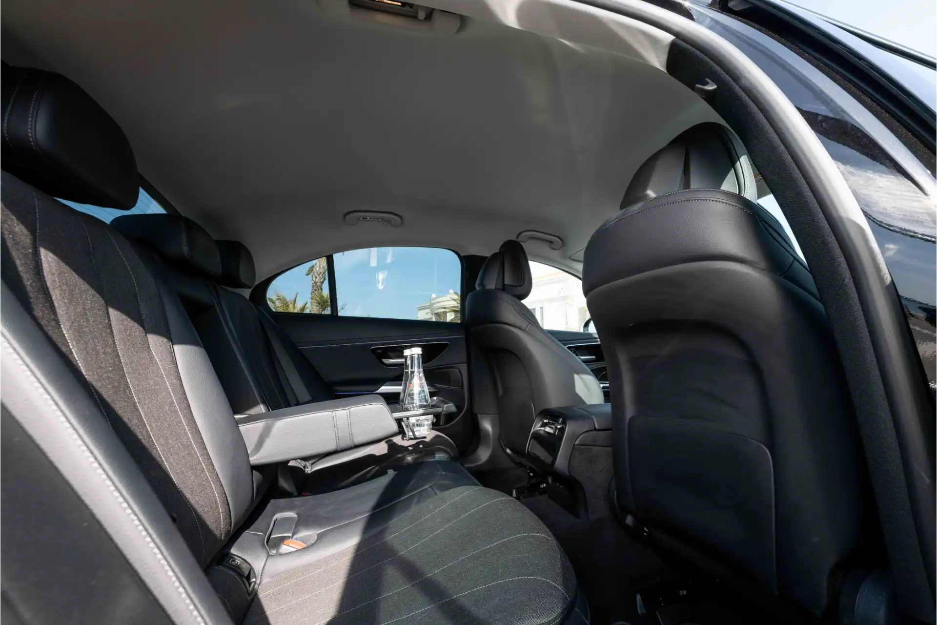 Rear interior of a Mercedes E-Class. Black leather bench seat, center armrest lowered with cup holder and bottle of water, comfortable legroom, understated finishes.