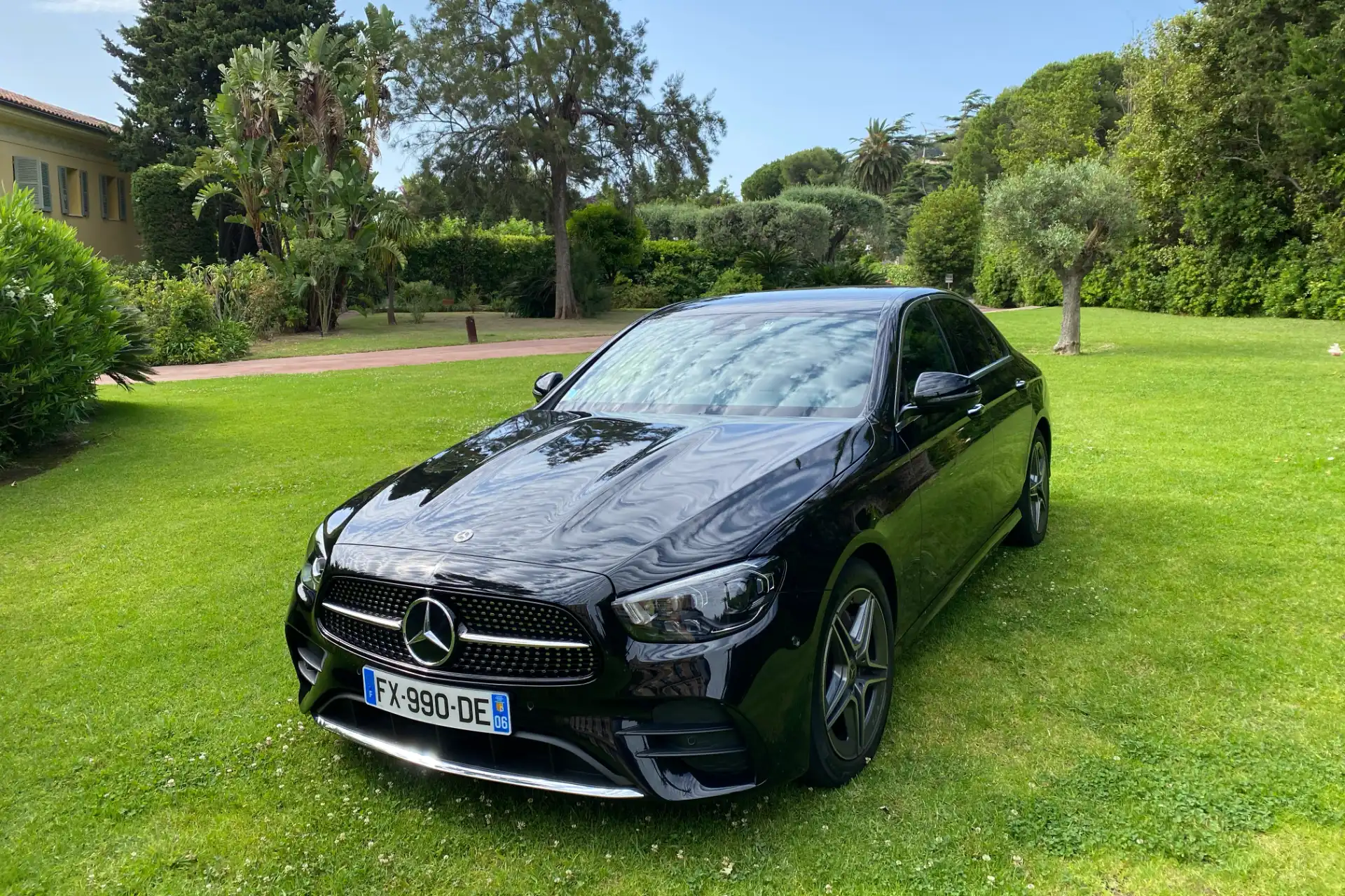 Black Mercedes E-Class seen from the front at a slight angle, parked in a tree-lined garden. Shiny bodywork with reflections of the sky, five-spoke alloy wheels, green residential environment.