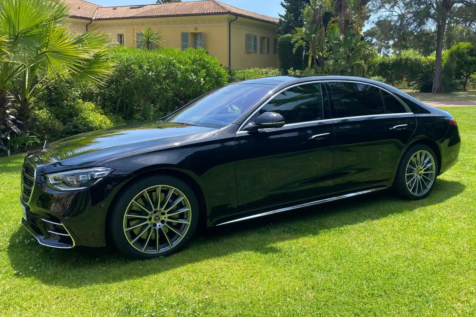 Black Mercedes S-Class sedan seen from the side, parked on a lawn in front of a villa surrounded by palm trees and dense vegetation. Multi-spoke alloy wheels, tinted windows, glossy bodywork.