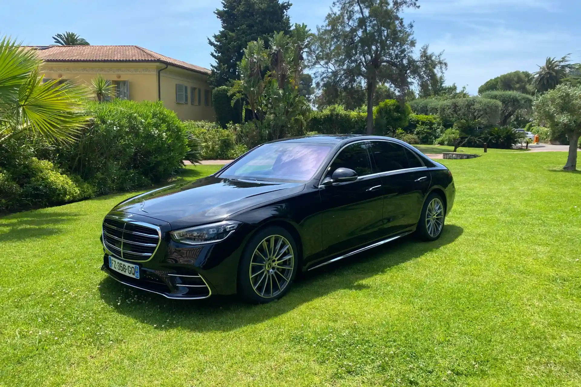 Black Mercedes S-Class seen from a front three-quarter angle, parked on a well-kept garden with a yellow house in the background. Large chrome grille, slim LED headlights, French license plate visible.