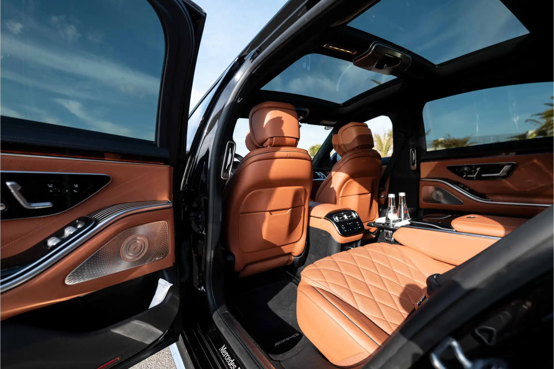 Rear interior of a Mercedes S-Class with doors open. Quilted brown leather seats, panoramic glass roof, rear center console with controls and bottles of water, high-end finishes.