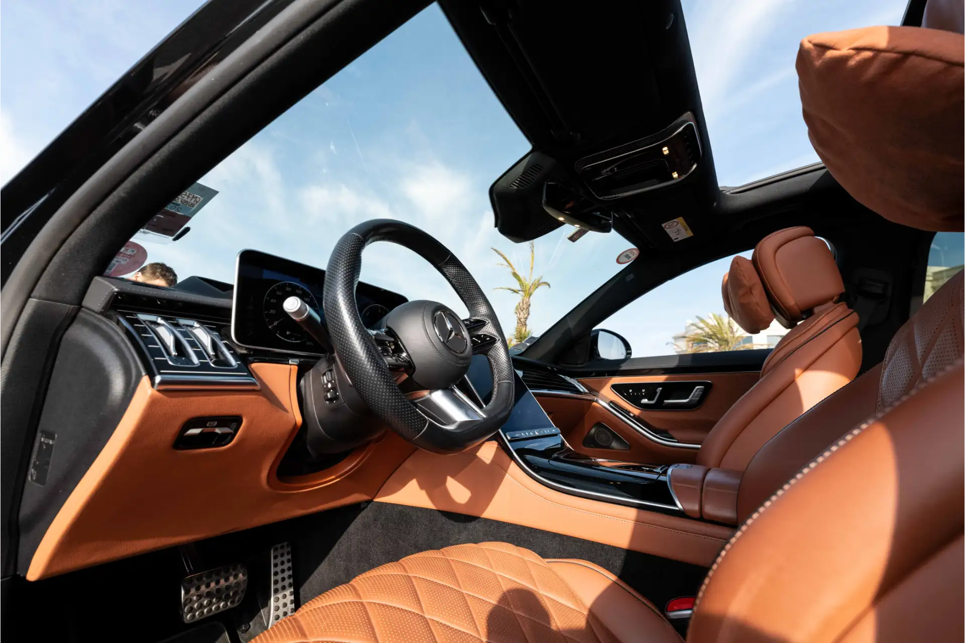 Driver’s cockpit of a Mercedes S-Class. Multifunction steering wheel with Mercedes logo, dual digital screens, glossy black inserts, and brown leather on the dashboard and seats.
