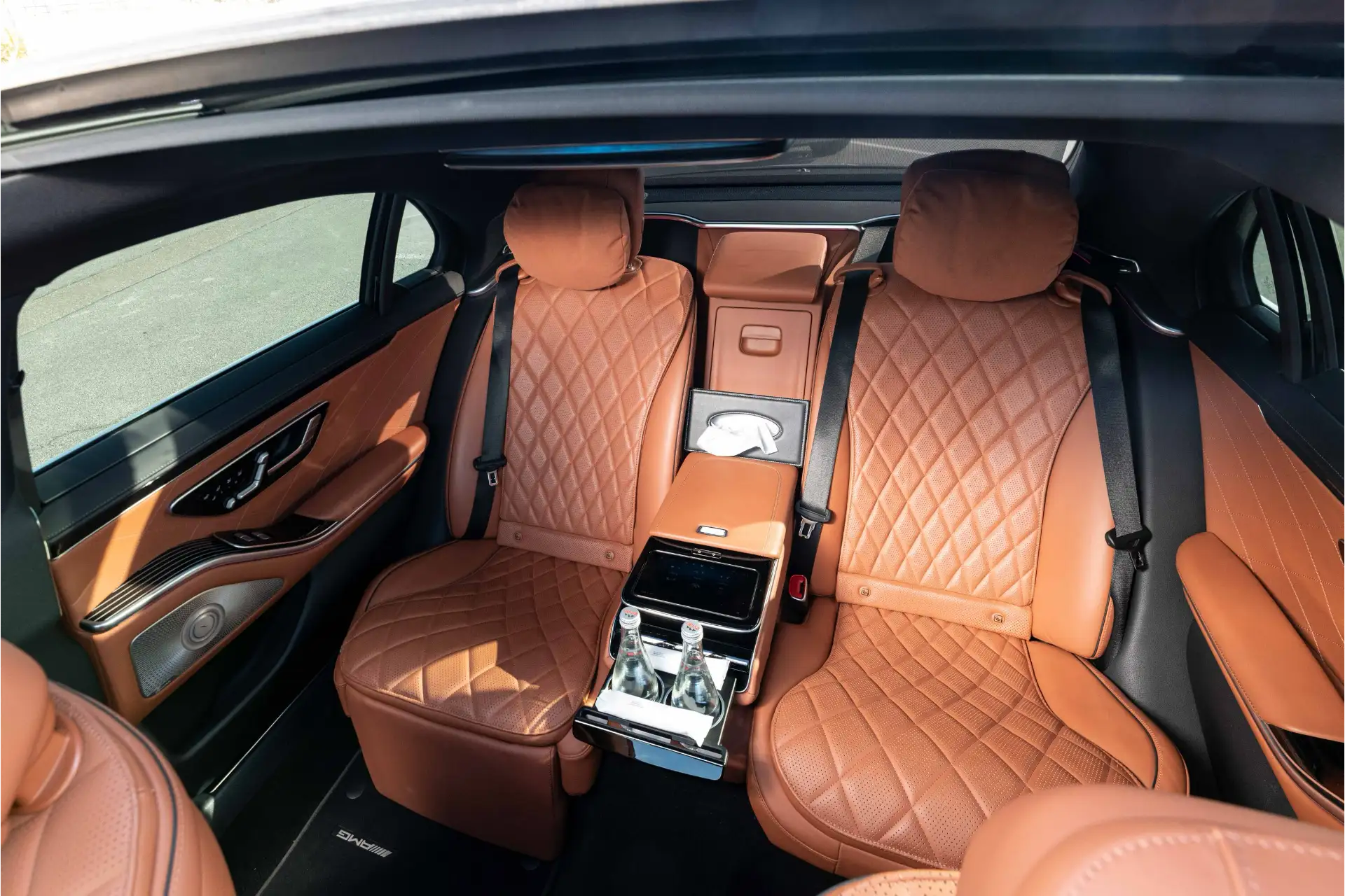 Rear bench of a Mercedes S-Class with quilted brown leather, two individual seat configuration with a central armrest, control screen, and compartment with bottles of water. Premium finishes and generous space.
