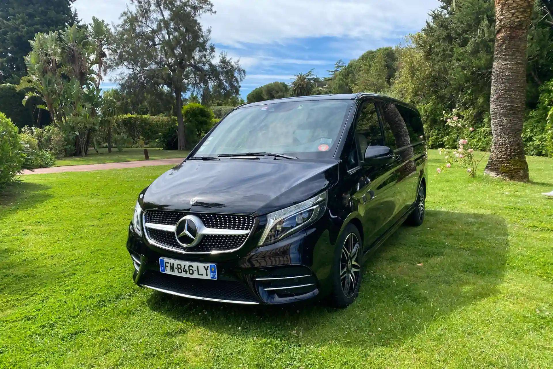 Black Mercedes V-Class seen from a front three-quarter angle, parked on a lawn in a tree-lined garden, black alloy wheels, chrome grille with central star.