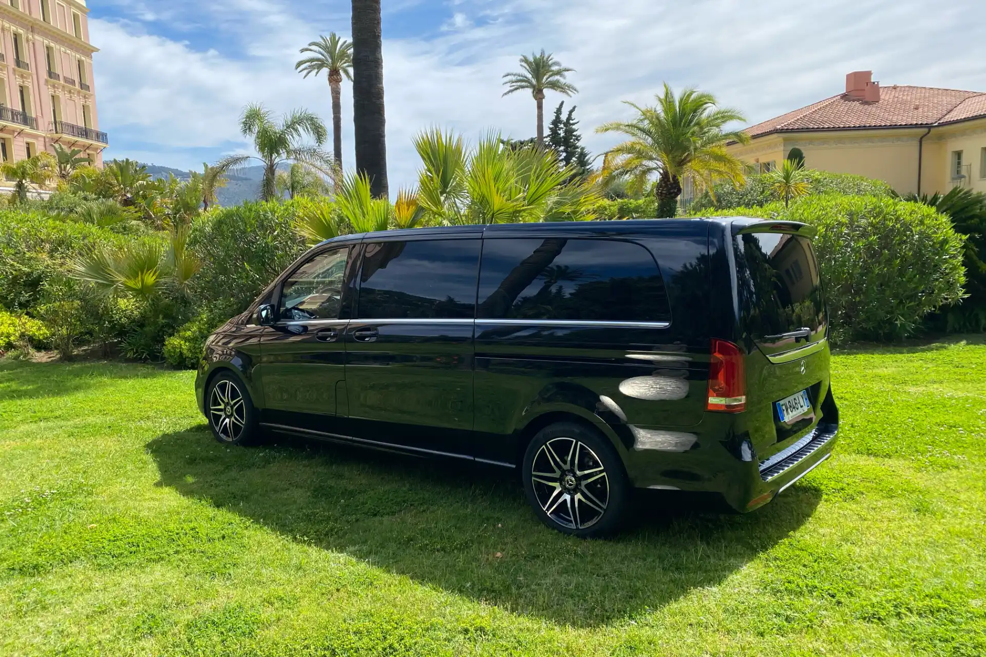 Black Mercedes V-Class seen from the rear left side profile, tinted windows, vertical rear lights, parked on a lawn with palm trees and a building in the background.