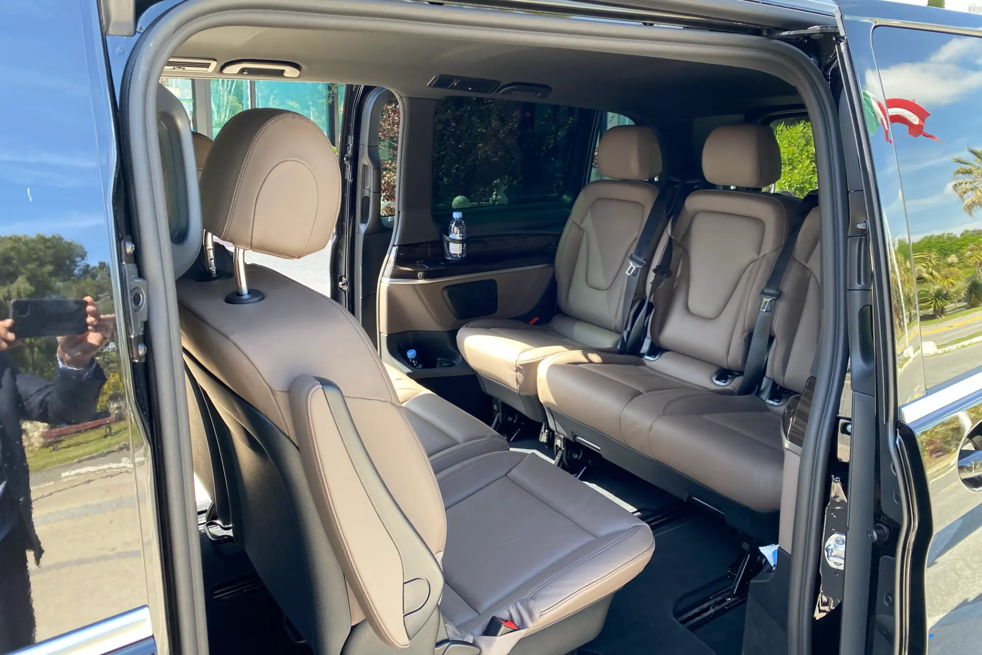 Interior of a Mercedes V-Class, side door open, beige leather seats, three-seat rear configuration, wood finishes, spacious interior.