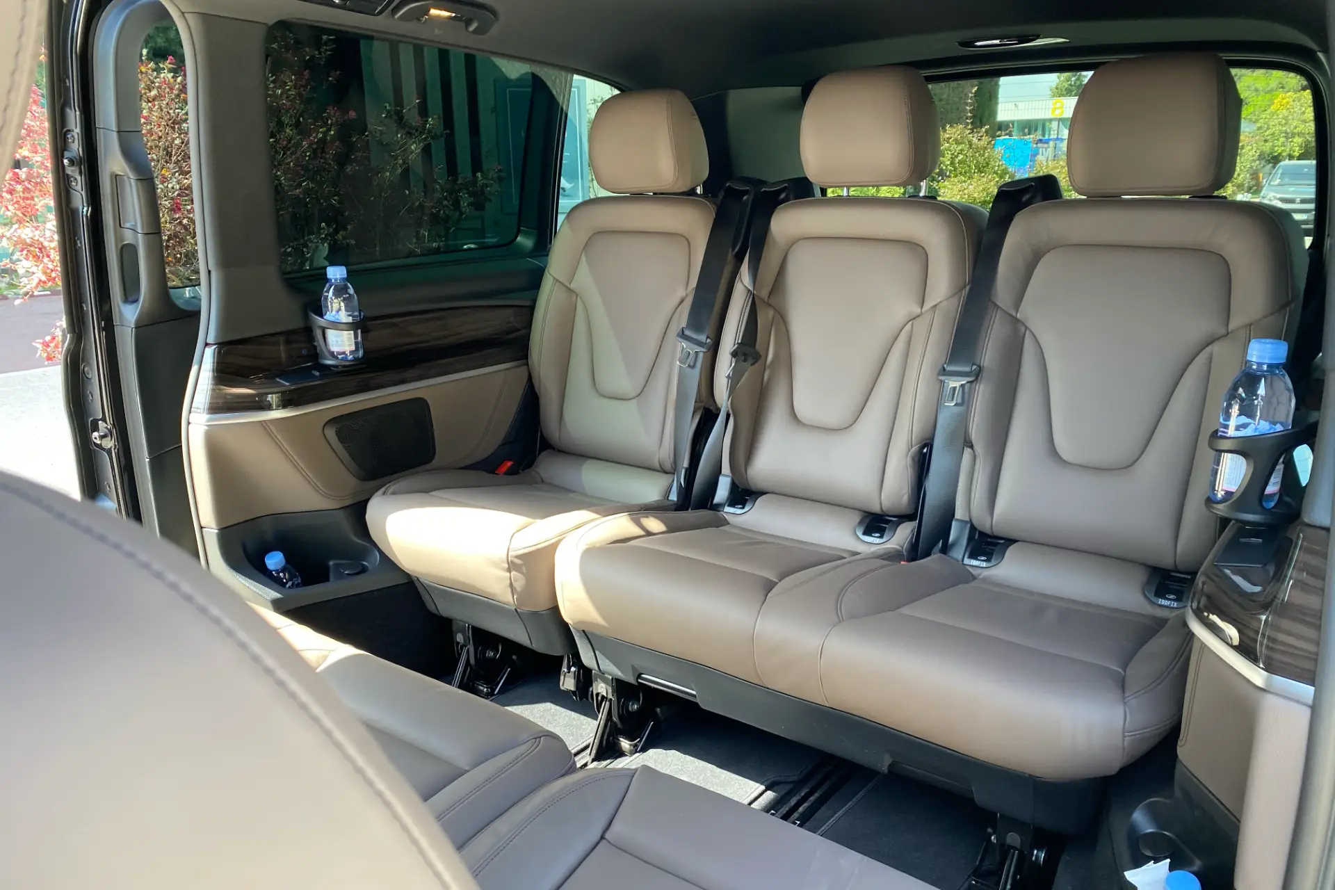 Rear row of a Mercedes V-Class in beige leather, side armrests with cup holders and bottle of water, wood finishes, sliding door open.