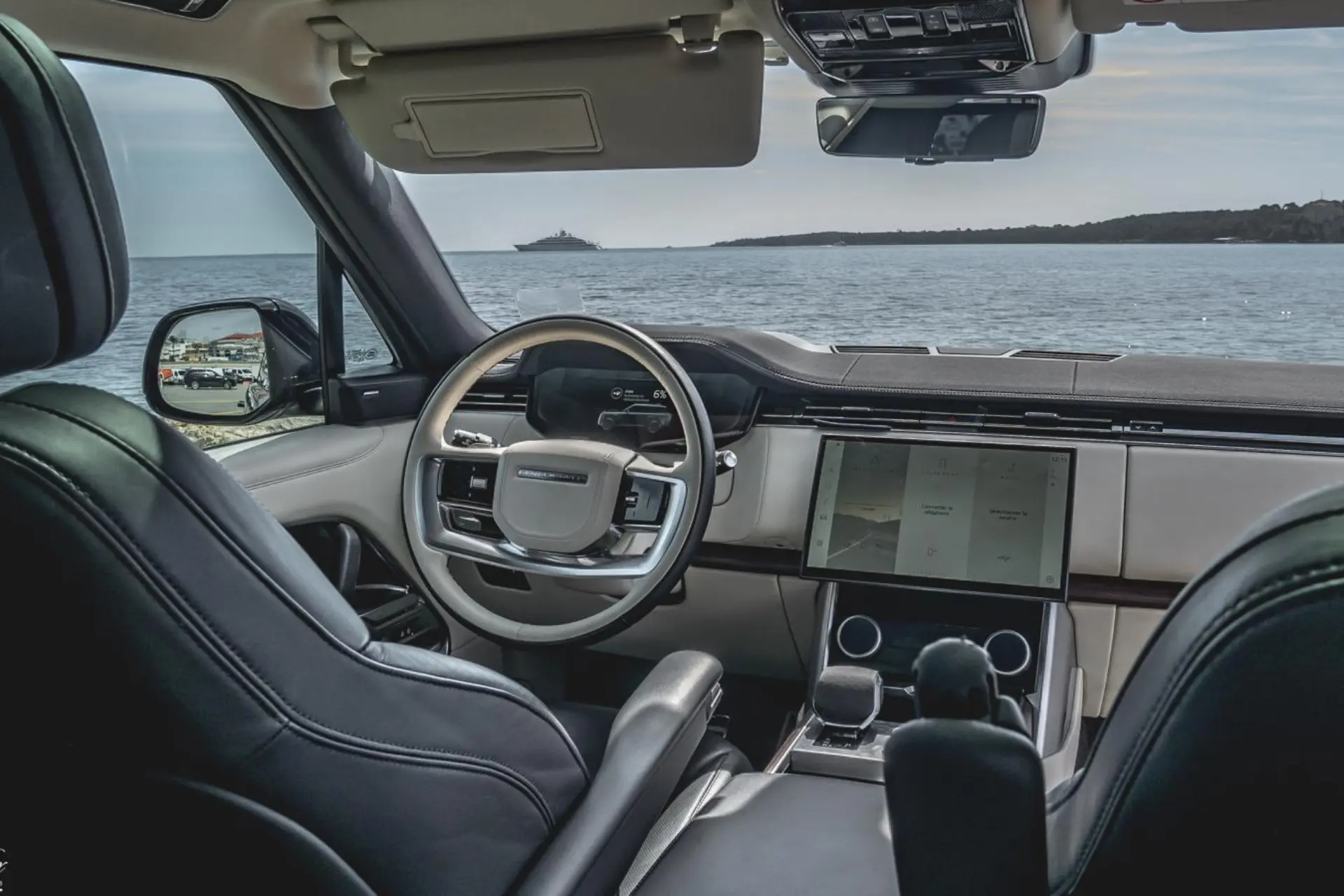 Front interior of a luxury SUV parked facing the sea. Light-colored steering wheel with a digital screen behind it. Large vertical central touchscreen. Two-tone dashboard with dark inserts. Through the windshield, the water and a yacht offshore are visible.