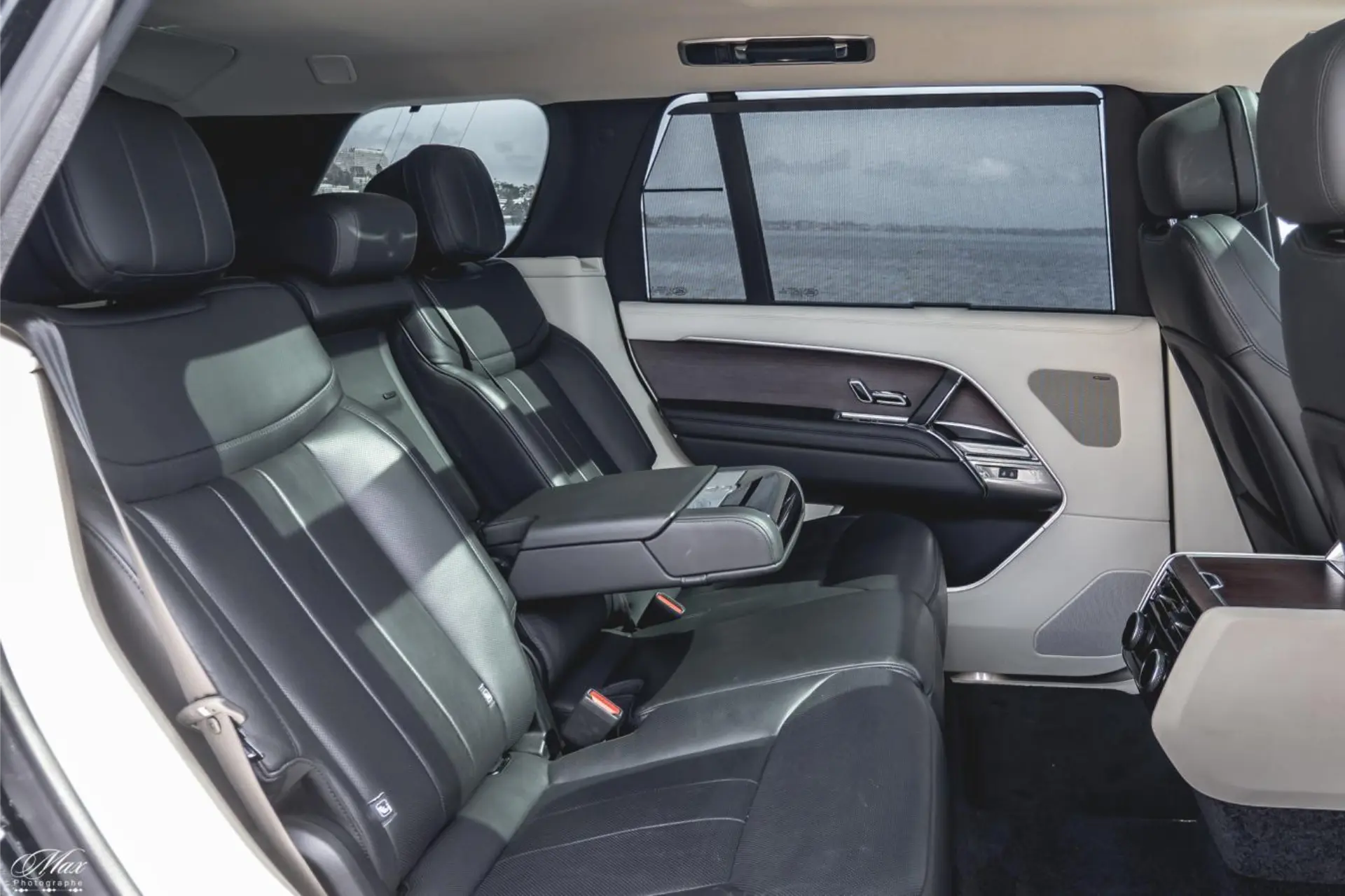 Rear bench in black leather with the central armrest lowered. Two-tone door panels with dark wood inserts and metal handles. Tinted windows with the sunshade raised. The sea is visible through the side window.