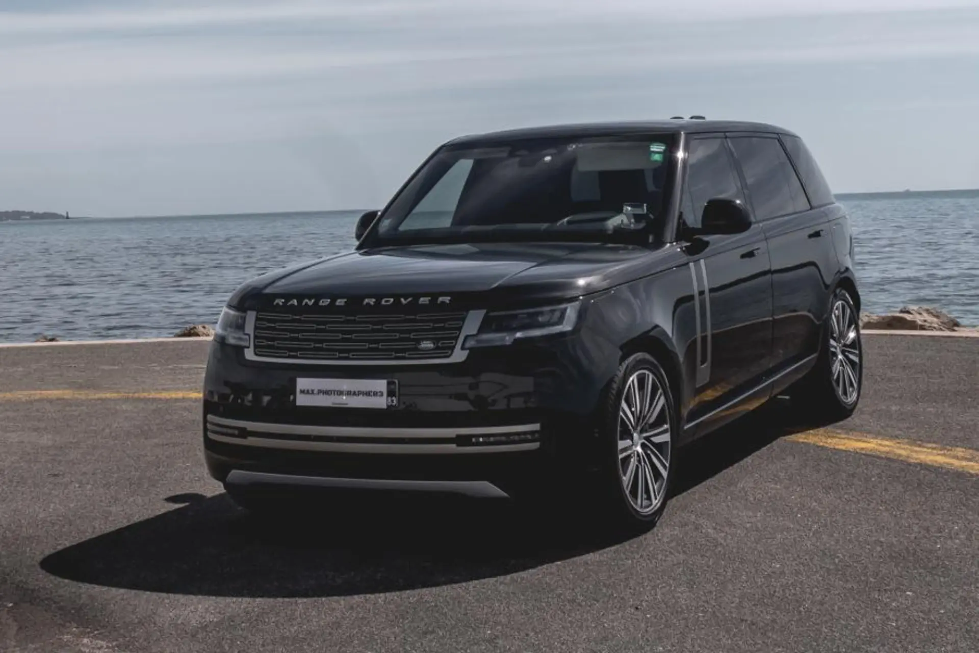 Black luxury SUV seen from a front three-quarter angle, parked on a seaside road. Wide grille and model name inscription on the front. Large multi-spoke wheels. Sea horizon in the background.