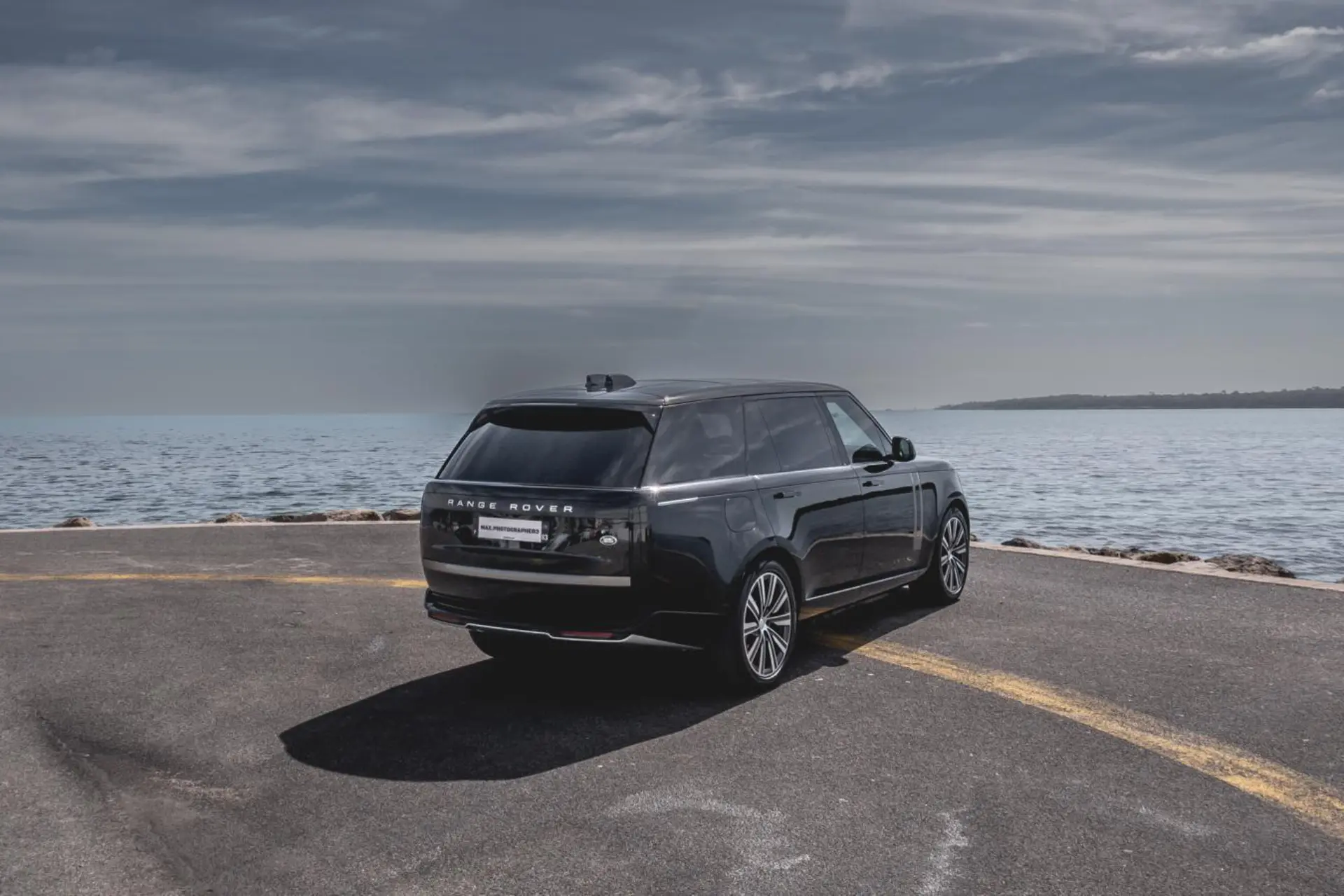 Black SUV seen from a rear three-quarter angle, parked near the water. Horizontal rear lights and tinted rear window. Straight roofline and imposing silhouette. Coastal road and sea in the background.