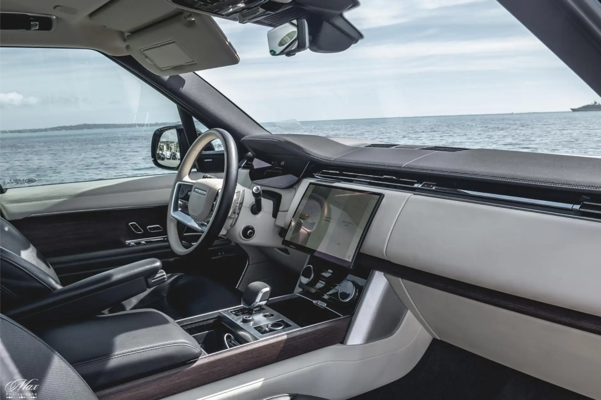 Front interior view from another angle. Light-colored steering wheel, vertical central touchscreen, and center console with gear selector. Dark wood finishes on the dashboard and door panels. View of the sea through the windshield.
