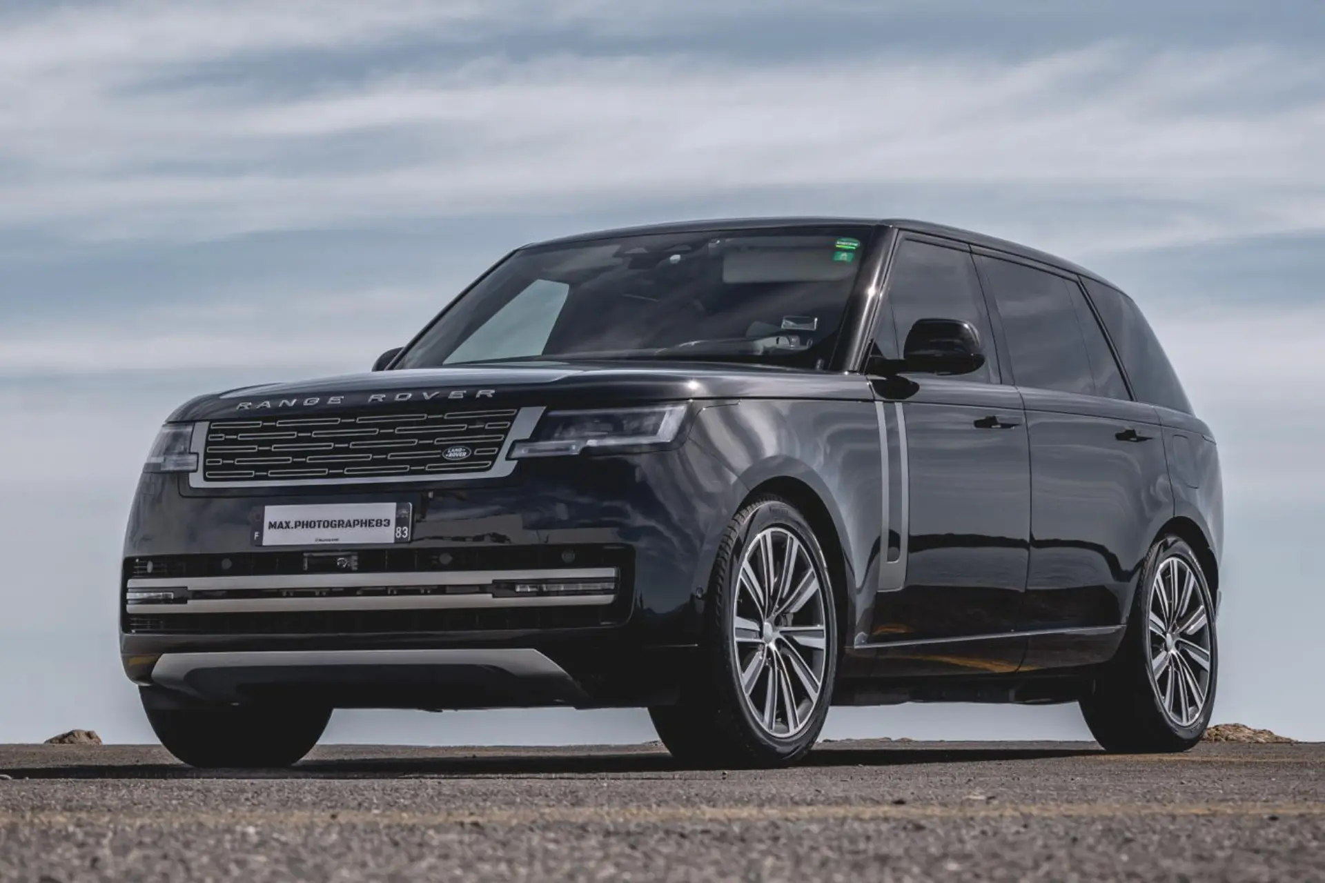 Black luxury SUV seen from the front in a slight low-angle view, parked on a seaside road. Wide grille with the model name on the hood. Rectangular headlights and multi-spoke alloy wheels. Cloudy sky and sea horizon in the background.