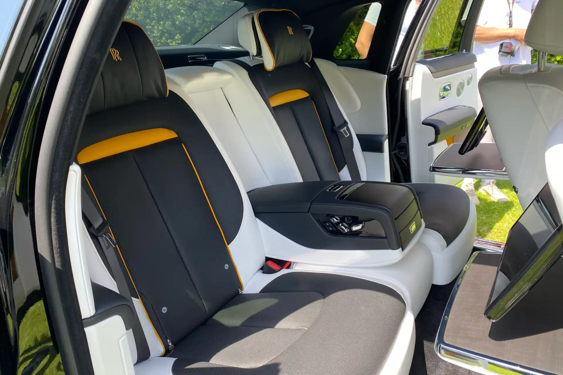 Rear interior of a Rolls-Royce with individual seats in two-tone black and white leather, yellow stitching and piping. Central armrest with integrated controls, high-end finishes.