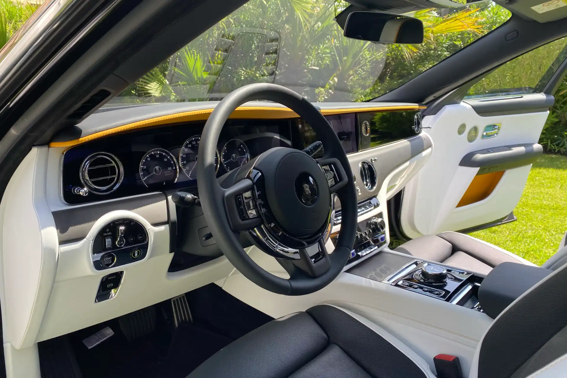 Driver’s cockpit of a Rolls-Royce. Steering wheel with RR logo, analog instrumentation, wood inserts and white leather with a yellow strip on the dashboard.