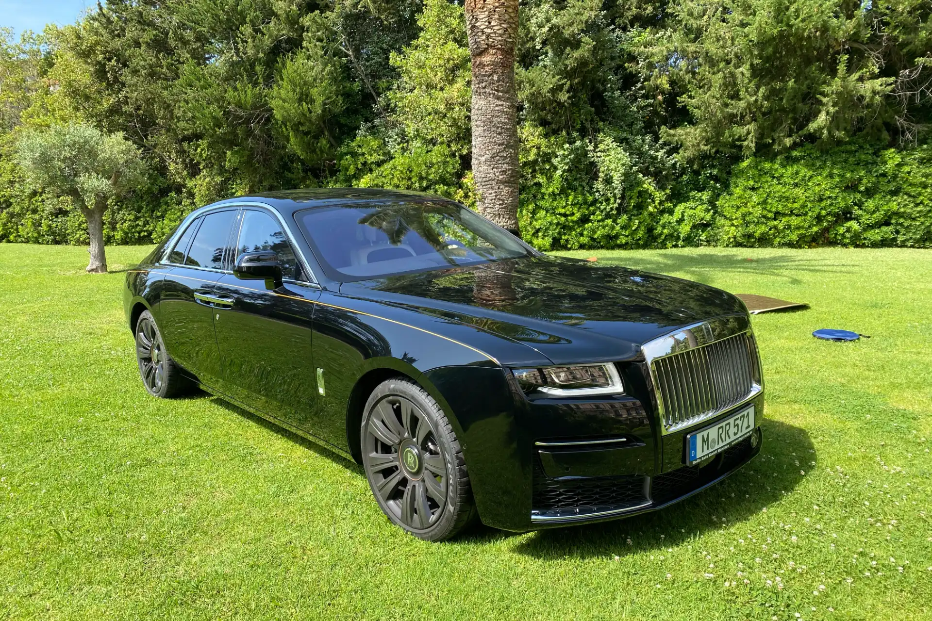 Black Rolls-Royce seen from a front three-quarter angle, parked on a tree-lined lawn. Large vertical chrome grille, black wheels, glossy bodywork.