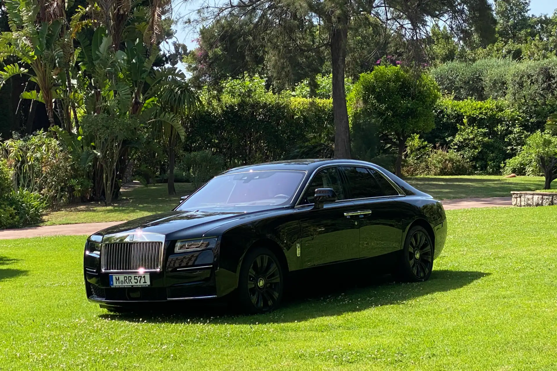 Black Rolls-Royce seen from the front left three-quarter angle, parked on a lawn with dense vegetation in the background. Elegant lines, tinted windows, black wheels.