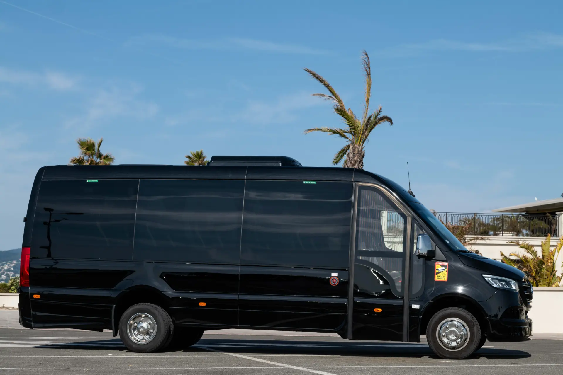 Large black van seen from the side, parked in a seaside parking area. Long body with tinted side windows. Palm trees and clear sky in the background. Raised roof and visible metal wheels.