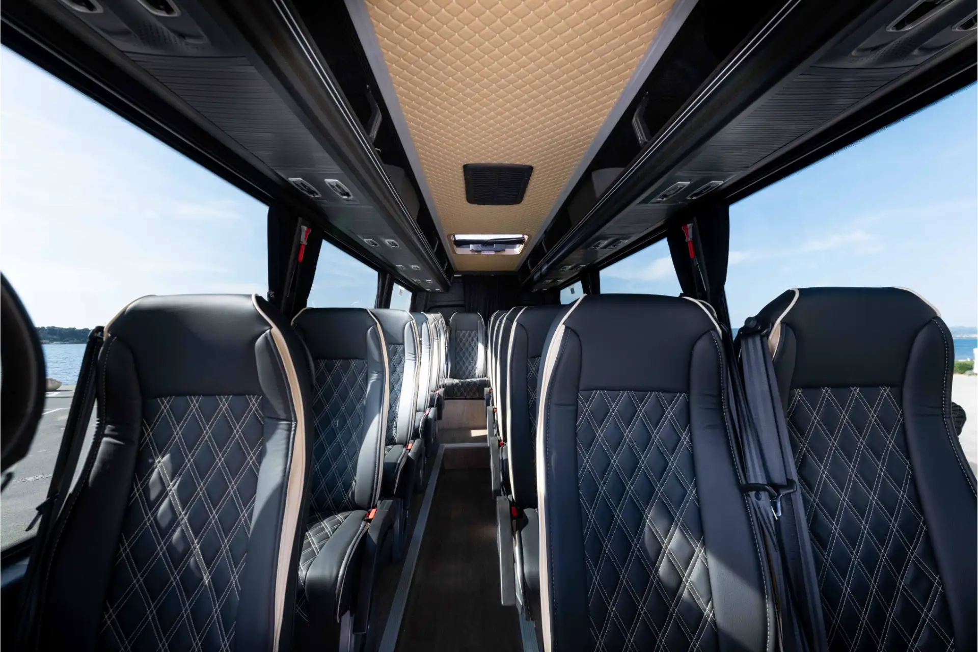 Interior of the vehicle with two rows of individual seats in black leather with diamond stitching. Clear central aisle. Light-colored ceiling with integrated lighting. Large side windows showing the sea.