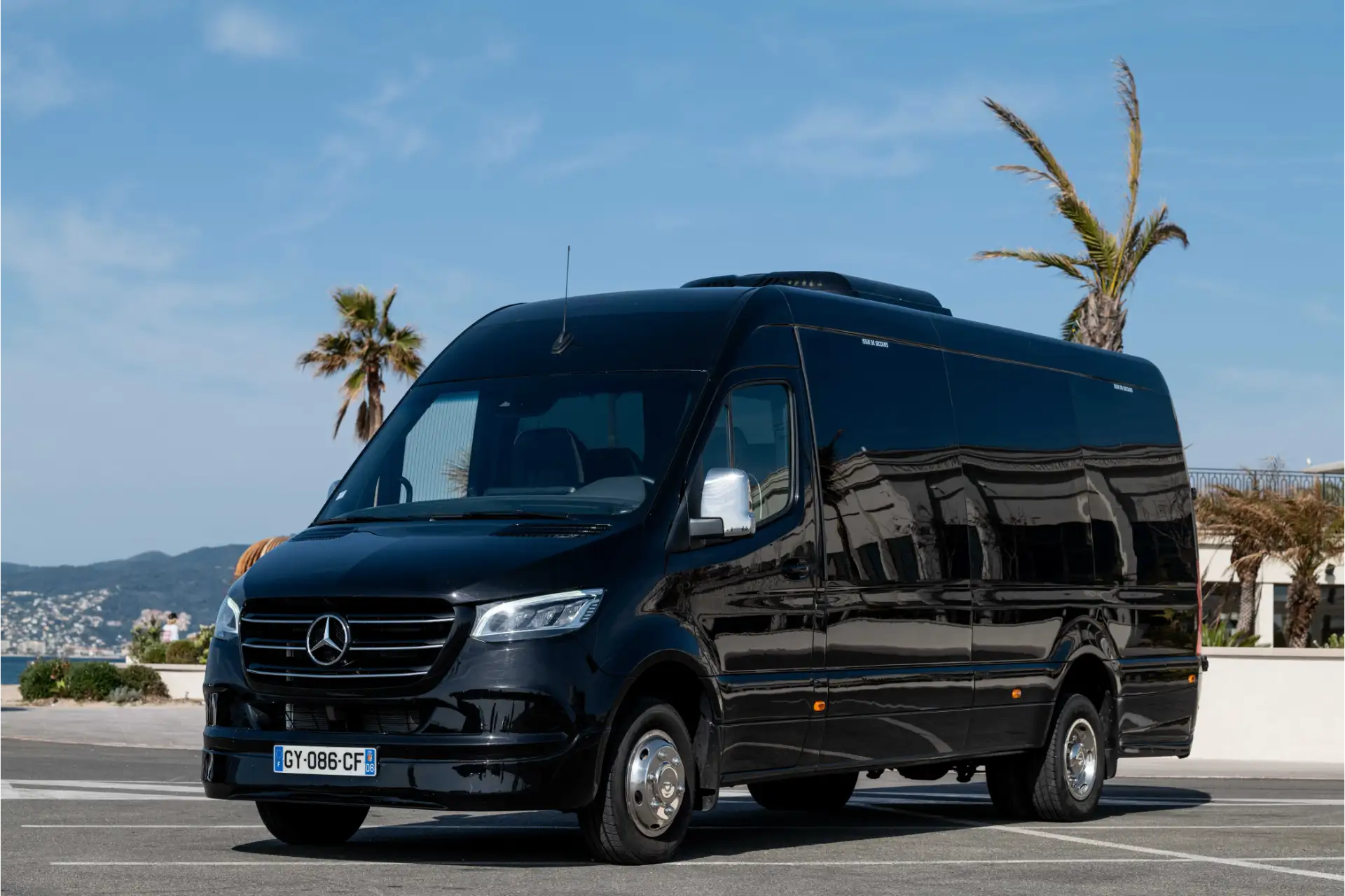 Black van seen from a front three-quarter angle, parked near the coastline. Grille with central logo, wide front headlights and large bumper. Palm trees, sea and buildings in the background.