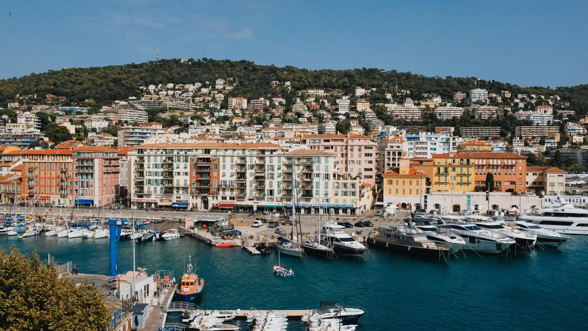 datacloud-congress Vue panoramique d’un port méditerranéen avec des yachts amarrés, des immeubles colorés en bord de mer et une colline boisée en arrière-plan.