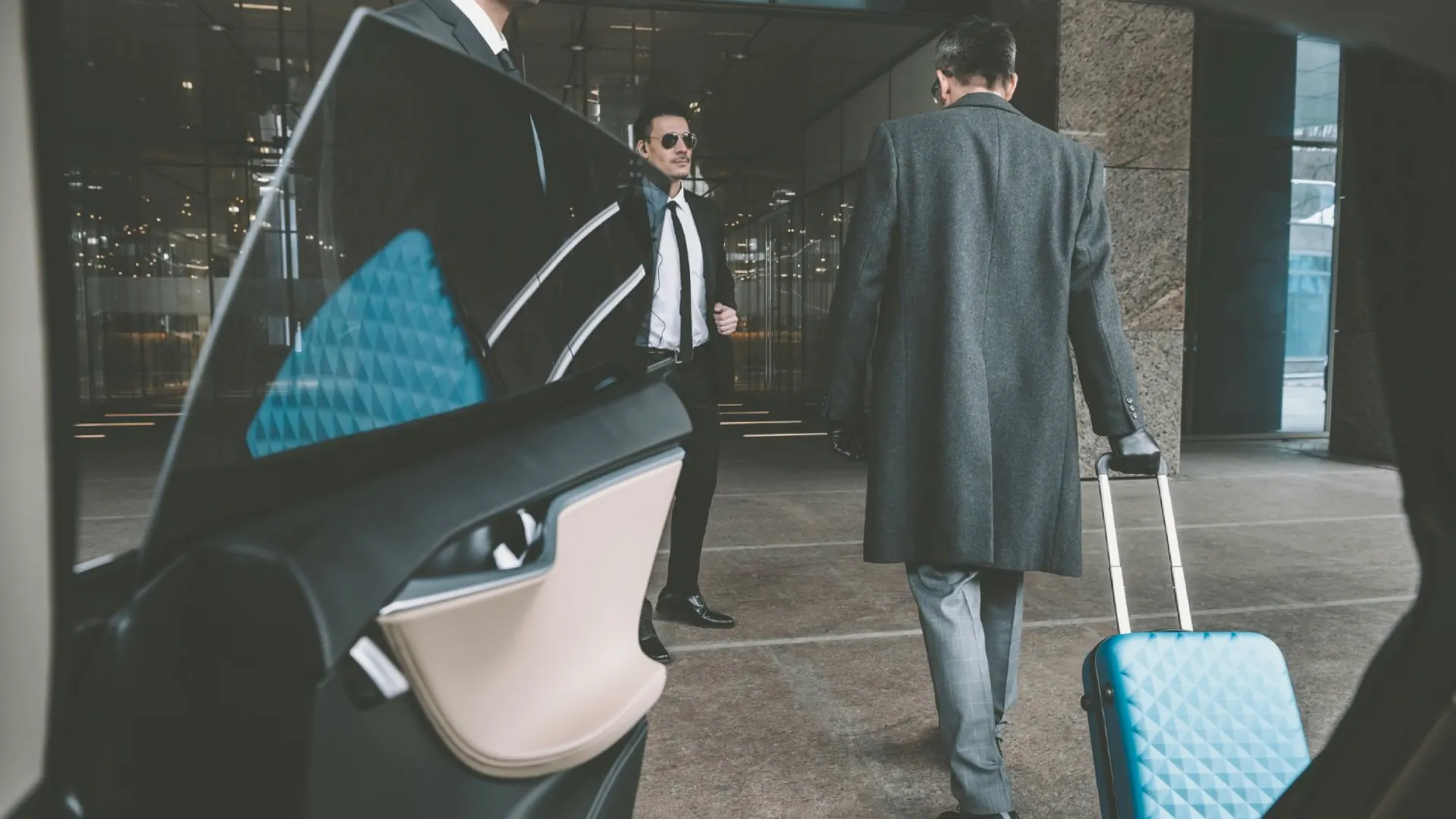 Premium chauffeur service for business travel, with a professional driver welcoming a suited client exiting a luxury vehicle in front of a modern building, corporate transfer with carry-on luggage.