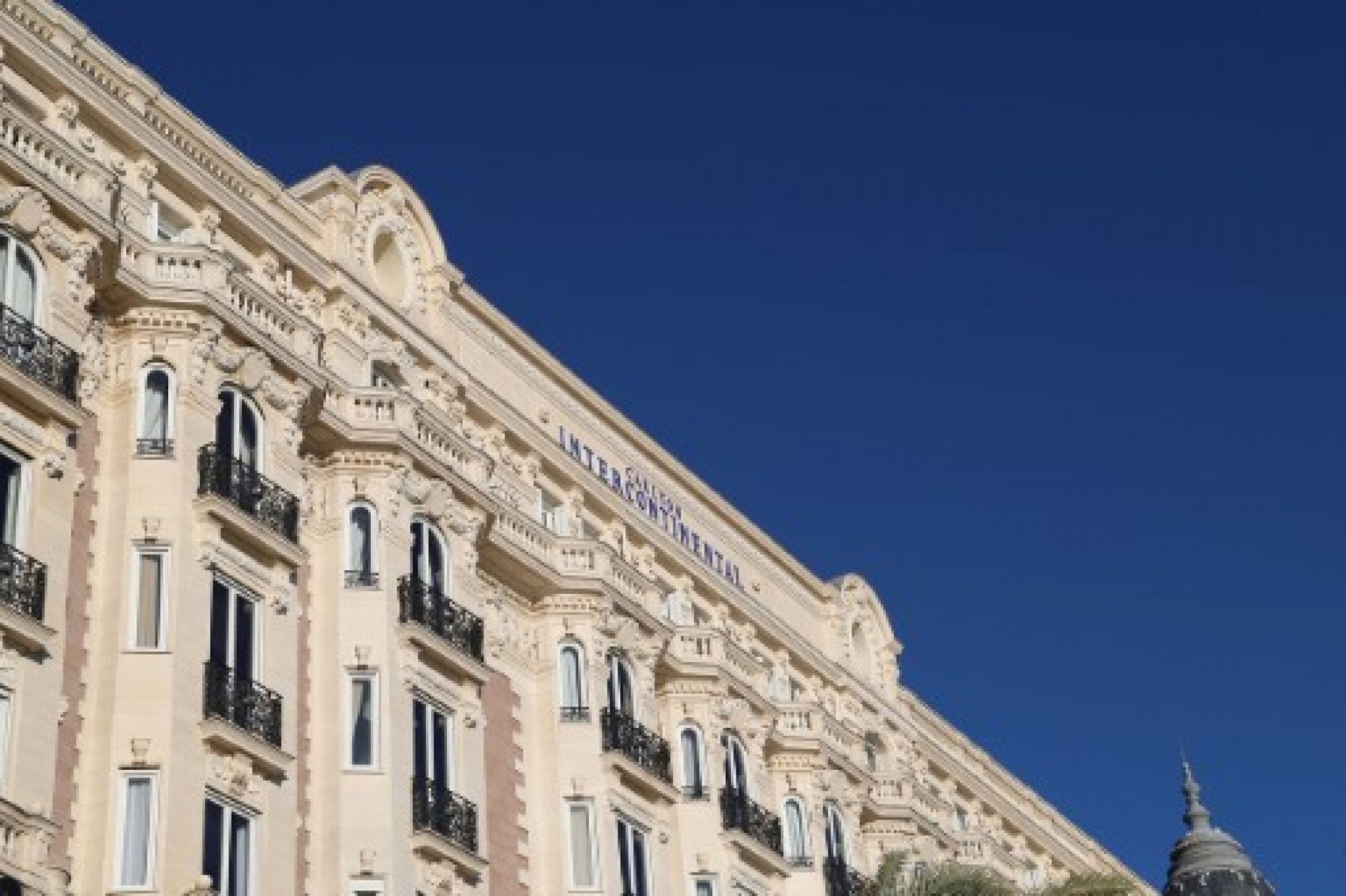 Façade élégante de l’hôtel InterContinental Carlton à Cannes, bâtiment historique de style Belle Époque avec balcons en fer forgé et détails architecturaux ornés, sous un ciel bleu dégagé.