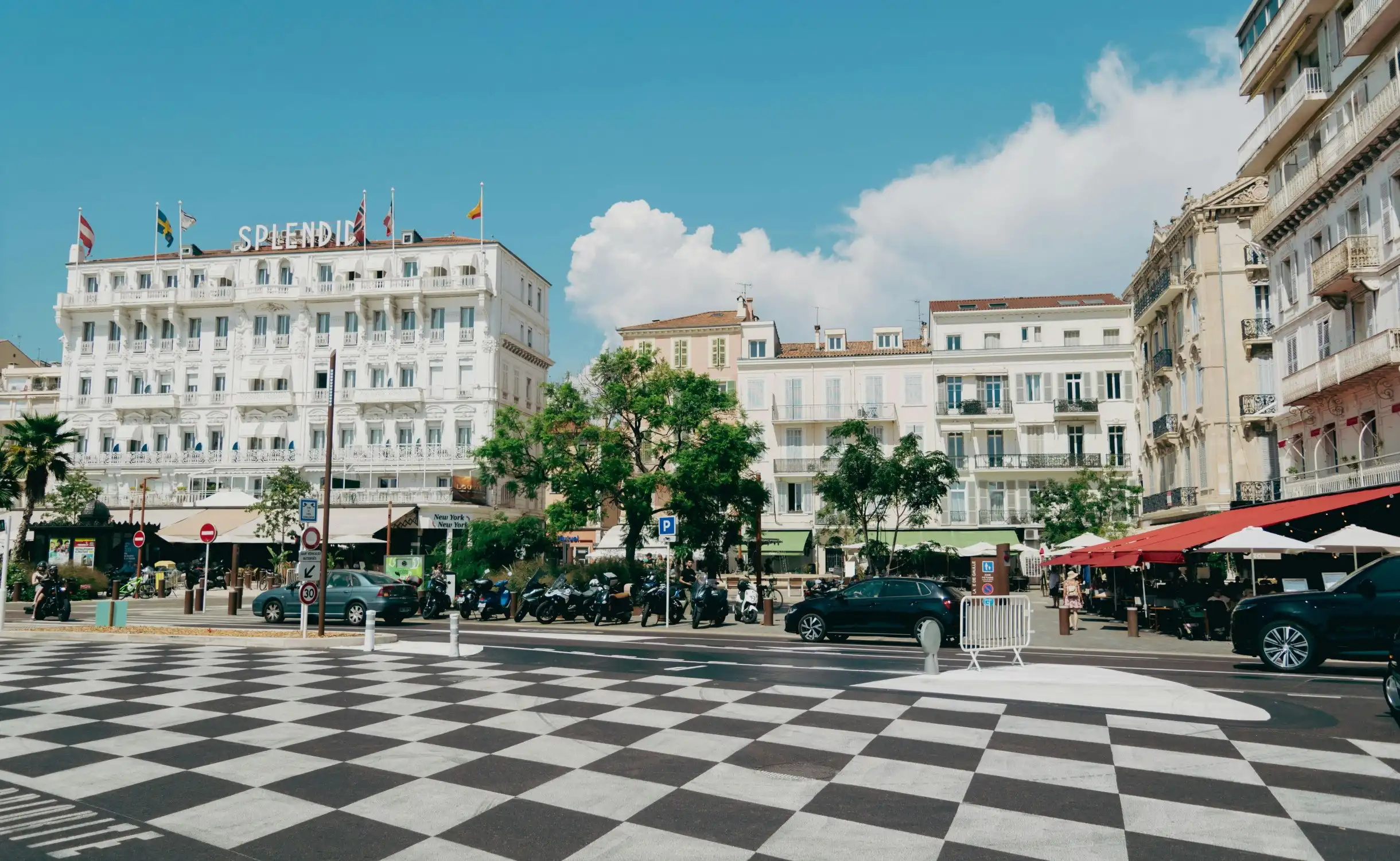 Place animée à Cannes avec l’hôtel Splendid en arrière-plan, façades typiques, terrasses de cafés, voitures et scooters, sous un ciel bleu sur la Côte d’Azur.