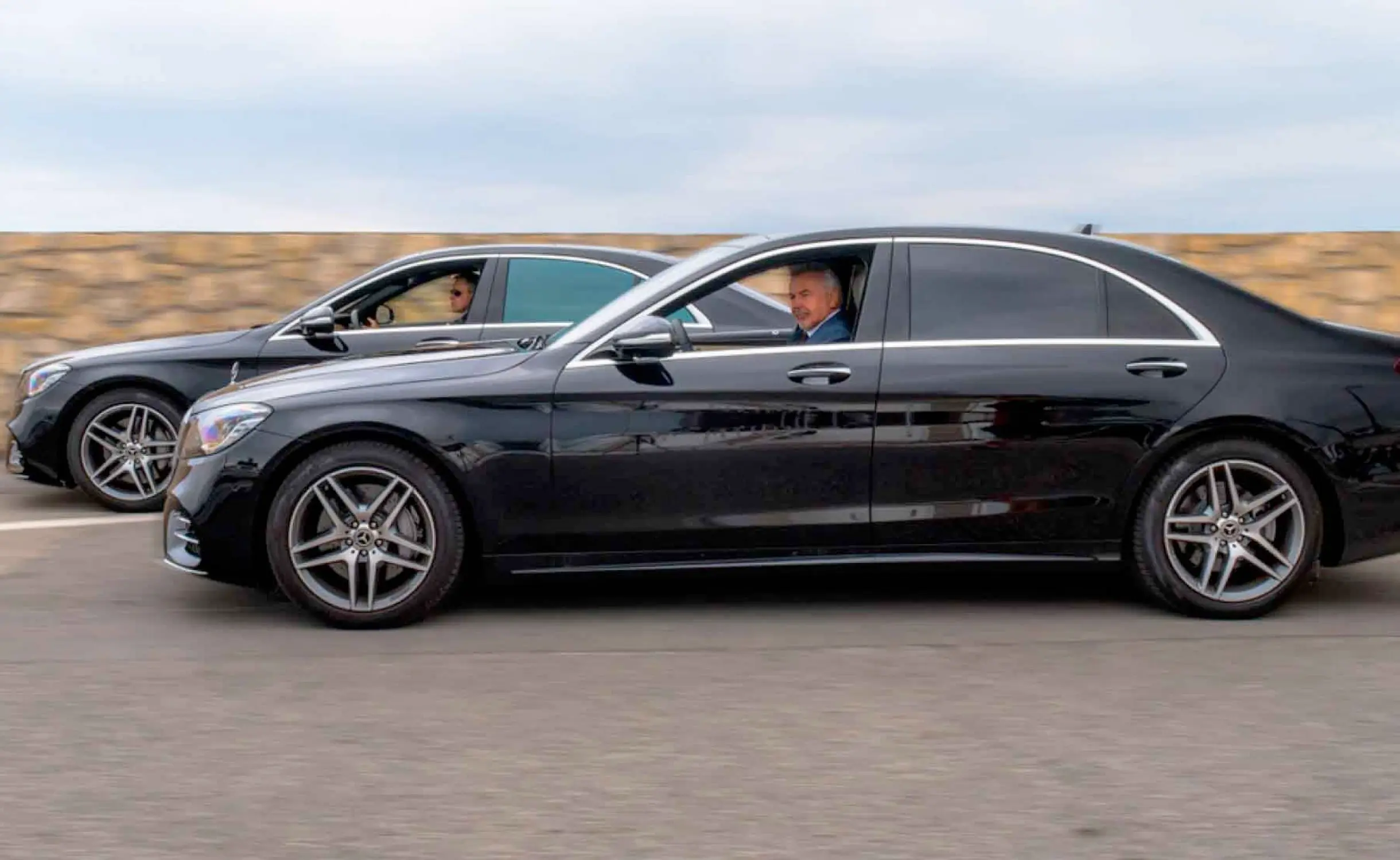 Deux berlines noires de luxe roulant côte à côte sur route, chauffeurs en costume visibles à l’intérieur, illustrant un service de transport privé haut de gamme.