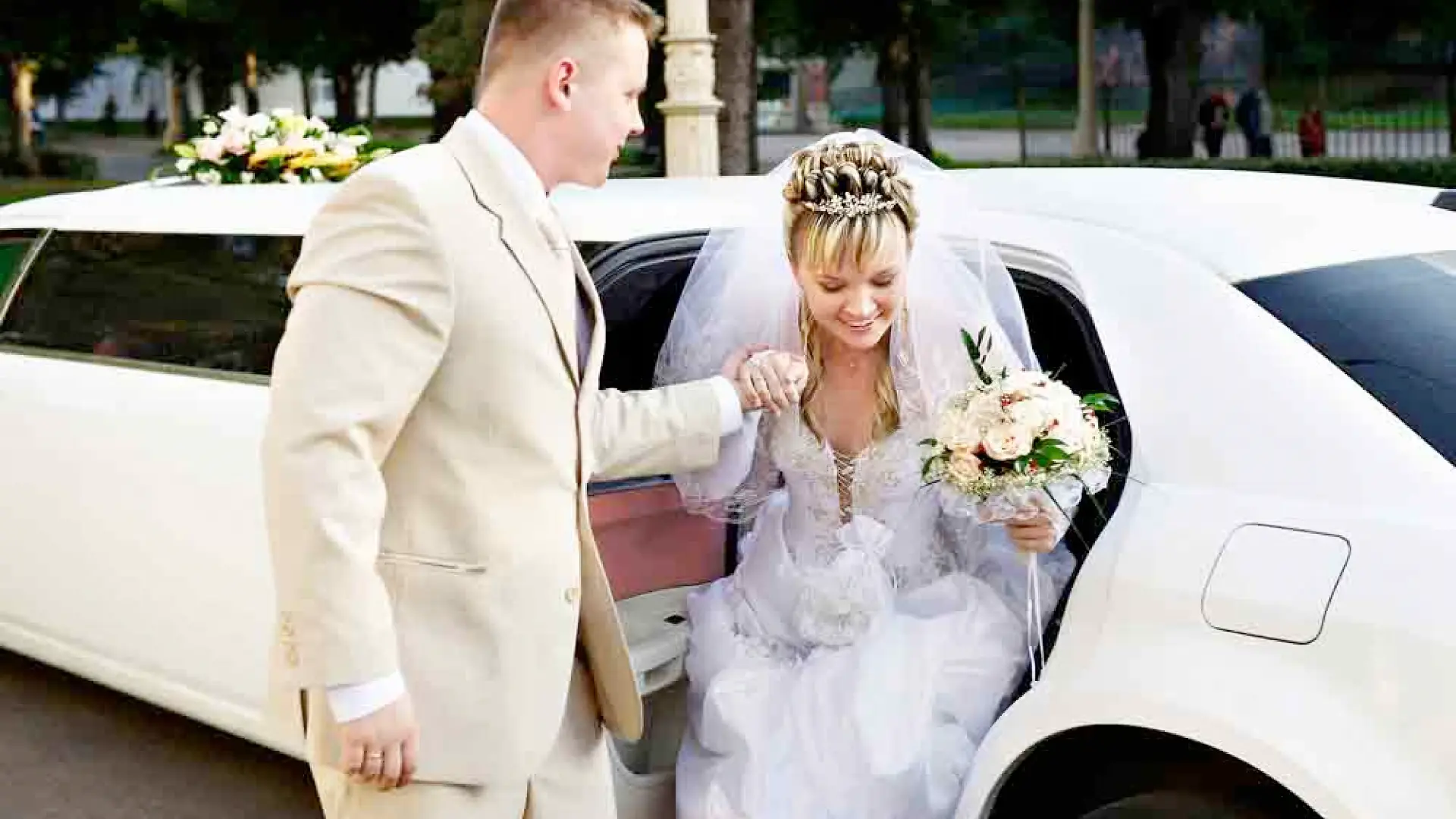 Bride in a white wedding dress and veil stepping out of a limousine, assisted by the groom in a beige suit, holding a bouquet during an outdoor wedding ceremony.